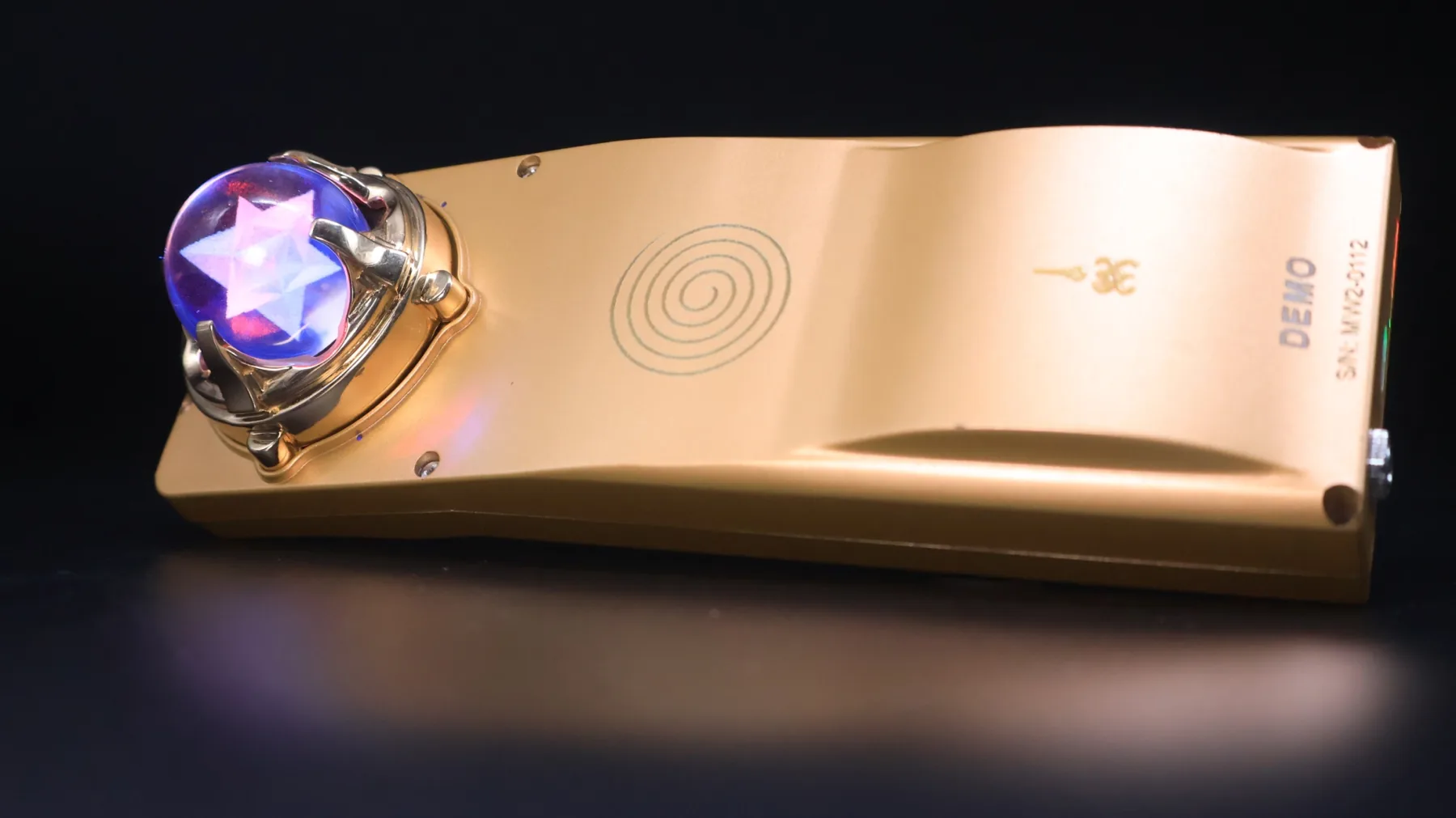 Gold Mandala Wand wellness device with violet illuminated quartz sphere on a dark reflective surface